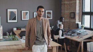 İki ırklı genç mimari bürosunun mimarlık bürosu ofis masasına yaslanıp kameraya bakıyor.