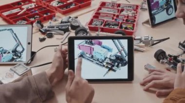 Masada oturmuş dijital tablet ekrandaki robot model görüntüsüne bakan ve detayları tartışan çocukların yüksek açılı dikiz görüntüsü