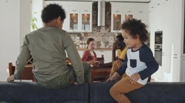 Orta uzunlukta iki Afro-Amerikan kardeş kanepenin arkasında emeklerken aileleri arka planda yemek masasında oturuyor.