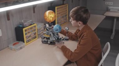 Modern beyaz çocuğun masada oturup okul projesi için robotik güneş sistemi modeli yaratırken yüksek açılı görüntüsü.