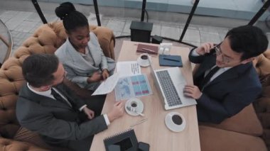 Asyalı erkek, beyaz erkek ve resmi takım elbiseli Afro-Amerikan kadın gündüz vakti çatıdaki restoranda oturmuş telefonla konuşuyor, bilgisayar ve belge kullanıyor.