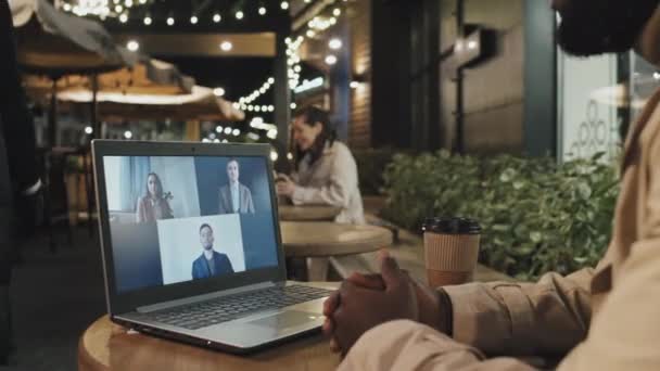 オンライン会議中に彼の外国人同僚と話すラップトップの前で夜遅くに屋外に座っている現代アフリカ系アメリカ人男性 — ストック動画