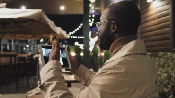 アフリカ系アメリカ人の男性は 夜遅くに屋外のカフェテーブルに座って デジタルタブレットを使用してオンライン会議中に彼の外国人同僚と話す — ストック動画