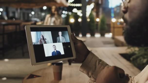 アフリカ系アメリカ人の男性が夜遅くにカフェのテーブルに座って デジタルタブレットを使用してオンライン会議中に彼の外国人同僚にアイデアを共有する — ストック動画