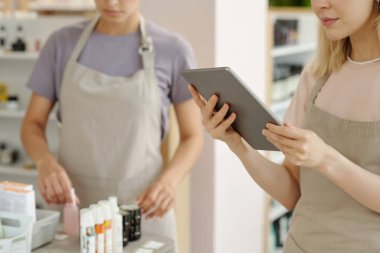 Kozmetik süpermarketinde çalışırken çevrimiçi güzellik ürünleri çeşitliliğine bakan önlüklü mağaza asistanının elleri