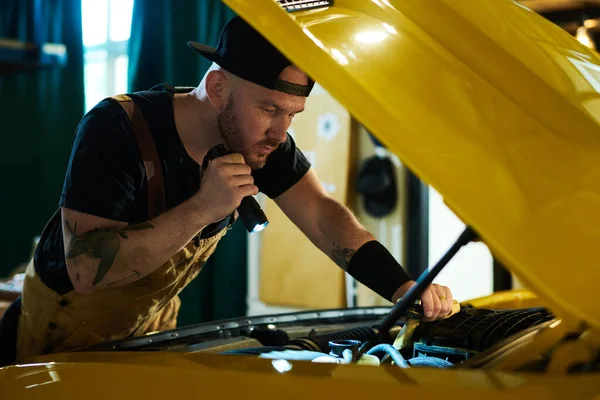 自動車修理工場の若い整備士は そのモーターに懐中電灯を指示しながら 黄色の自動車のエンジンをチェック — ストック写真