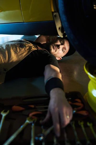 年轻的修理工躺在车底用工具从开着的工具箱或箱子里取出扳手 修理零件 — 图库照片