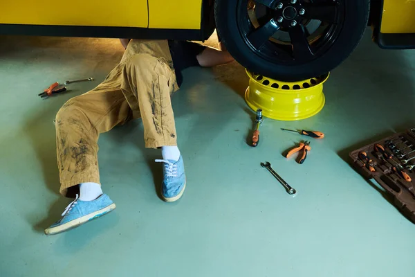 Nogi Młodego Mechanika Lub Mechanika Kombinezonach Leżących Pod Żółtym Samochodem — Zdjęcie stockowe