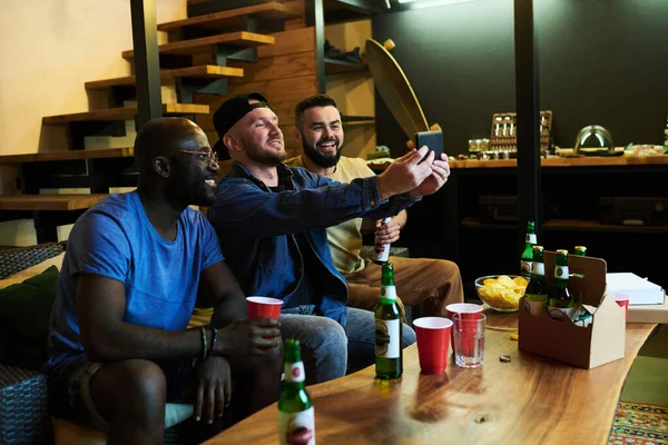 三位快乐的跨文化好友一边在智能手机上自作主张 一边在餐桌前的沙发上放松一下 一边喝啤酒和吃零食 — 图库照片