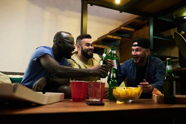 在庆祝胜利的同时 年轻快乐的不同种族的朋友和足球迷们在餐桌边喝着啤酒 喝着杯子 吃着点心 叮当作响 — 图库照片
