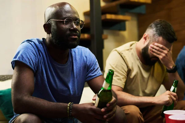 年轻而心烦意乱的非裔美国人 拿着一瓶啤酒和他的朋友一起在车库里看电视转播足球赛 — 图库照片