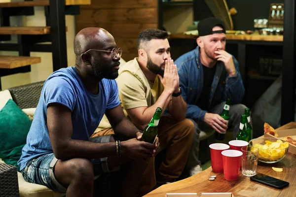 三个跨文化的年轻男性朋友一边看电视 一边在桌前放啤酒和零食 表达了他们的担忧 — 图库照片