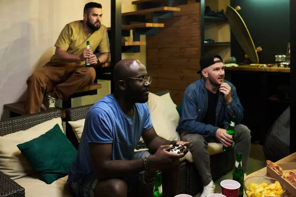 二人の若いです男とともにビールを見テレビ画面とビデオゲームを見ながら黒男とともにジョイスティック座ってボタンを押す — ストック写真