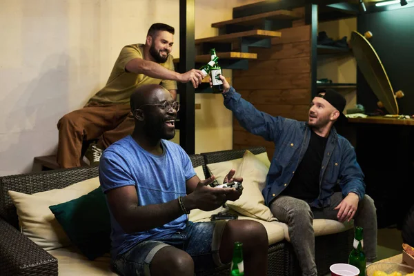 两个男人一边喝着啤酒一边和他们的非洲裔美国朋友坐在沙发上玩电子游戏 — 图库照片
