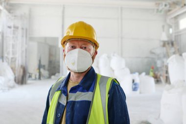 Modern fabrika işçisi ya da dağıtım deposu koruyucu solunum cihazı, iş giysisi ve güvenlik kaskı takmış kameraya bakıyor.