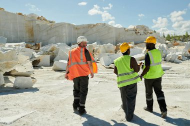 Modern fabrikanın bölgesinden geçen üç mermer ocağı işçisinin iş kıyafetleri ve güvenlik miğferlerinin arka görüntüsü.