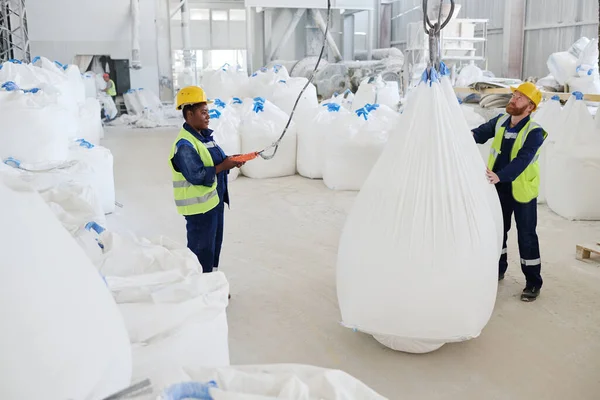 若い男性労働者を助ける彼の女性の同僚とリモートコントロールスイッチリフト巨大な重い袋で原材料でワークショップ — ストック写真