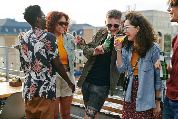 Group of young cheerful and affectionate friends enjoying outdoor party in rooftop cafe while having beer and cocktails and chatting