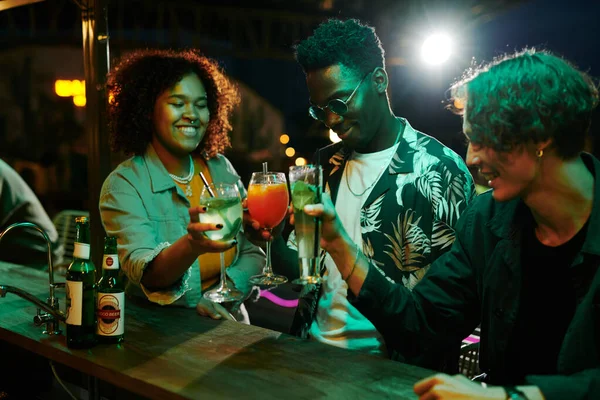 三个快乐的不同文化的年轻人在夜总会享受派对的时候 在柜台边喝着含酒精的鸡尾酒 — 图库照片