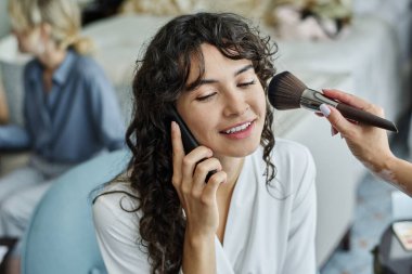 Makyajcı yanağına pudra sürerken, uzun dalgalı saçları olan mutlu bir gelin akıllı telefondan konuşuyor.