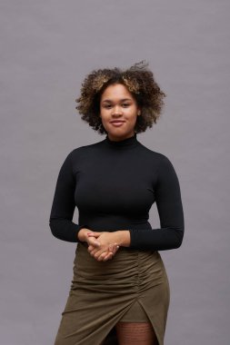 Gri arka planda tek başına dikilirken şık ve şık giyinmiş genç siyahi kadın kameraya bakıyor.