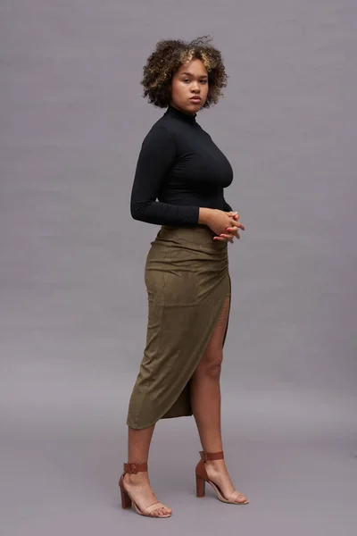 stock image Young elegant intercultural woman in black turtleneck, long brown skirt and high heeled sandals posing against grey background