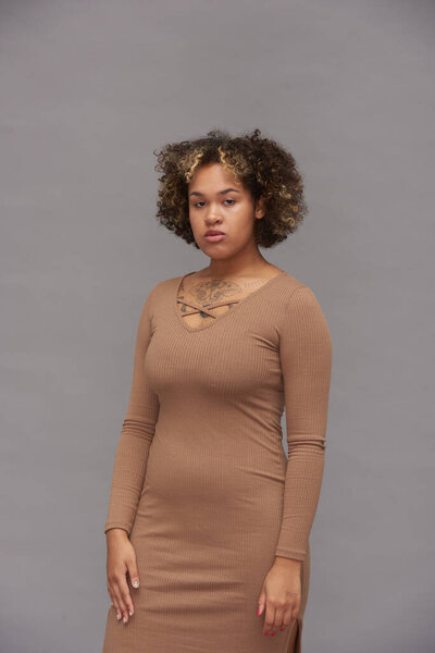 Young gorgeous woman in fashionable beige dress posing on grey background while standing in front of camera in isolation