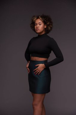 Stüdyoda kameranın önünde dikilirken gri arka planda poz veren şık giyimli genç, zarif bir bayan model.