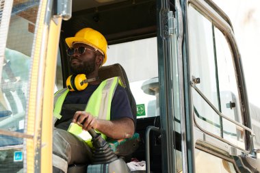 Genç Afrikalı Amerikalı inşaat kamyonu sürücüsü direksiyonun önünde makine kapısının önünde oturuyor ve iş sırasında onu sürüyor.