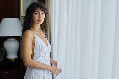Beyaz gelinlikli güzel esmer kadın pencerenin yanında şifon perdelerle kameraya bakıyor.