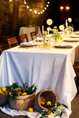 Beyaz masa örtüsüyle kaplı servis edilen şenlik masasının önü ve lüks restoranda yerde limon dolu üç sepet.