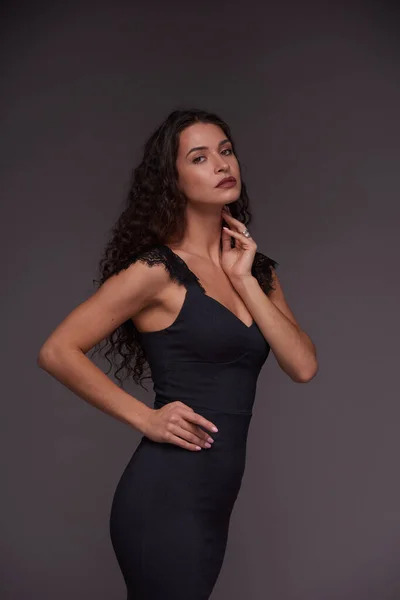スタジオでのフォトセッション中にグレーの背景の上にカメラの前に立つエレガントな黒の包帯のドレスの若いファッションモデル — ストック写真