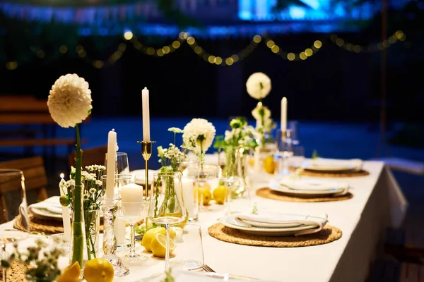 暗い青色の光に照らされたレストランでキャンドルや白いダリアの花で飾られた長いお祝いのテーブルの一部はロマンチックな雰囲気を作り出します — ストック写真