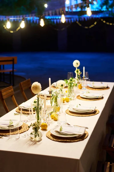 Long table covered with white tablecloth with dahlia flowers, candles, decorations and wineglasses surrounded by two rows of plates
