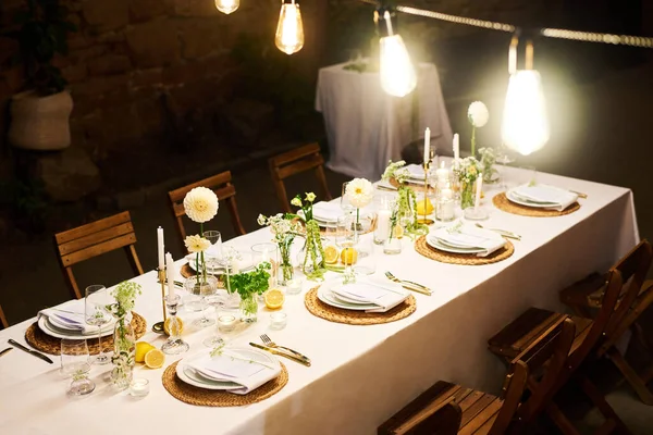 お祝いのテーブルの上に吊るされたいくつかの明るいランプは 結婚式のゲストと白いテーブルクロスに立っているオブジェクトを照らす — ストック写真