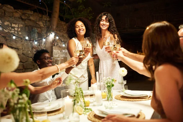 两个快乐的不同文化的年轻新娘和他们的婚礼来宾一起祝酒 并在喜庆的餐桌上用香槟酒轻轻摇曳着 — 图库照片