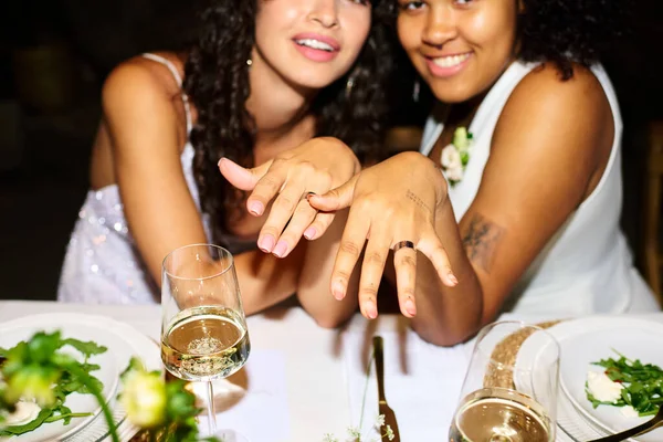 stock image Young intercultural lesbian couple showing their wedding rings on fingers while sitting by served table and holding hands in front of camera