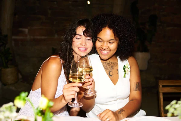 参加宴会时 穿着白色婚纱 头戴香槟酒长笛的年轻而快乐的跨种族女性坐在餐桌旁 — 图库照片