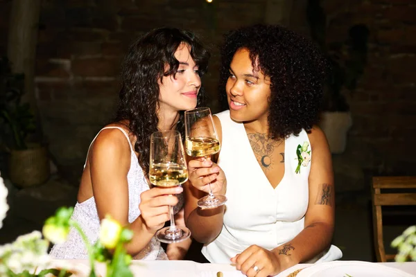 两个年轻的不同文化的新娘坐在婚宴餐桌旁 举杯祝酒的时候互相望着对方 喝着香槟 — 图库照片