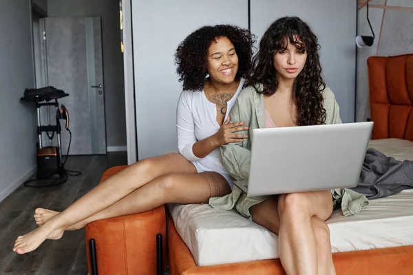 穿着白色家居服的快乐的年轻黑人女性坐在女友的床上 用笔记本电脑联网或视频聊天 — 图库照片