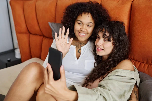 两个快乐的女朋友早上躺在床上休息 一边在电视上聊天 一边其中一个一边向自己的朋友挥手 — 图库照片