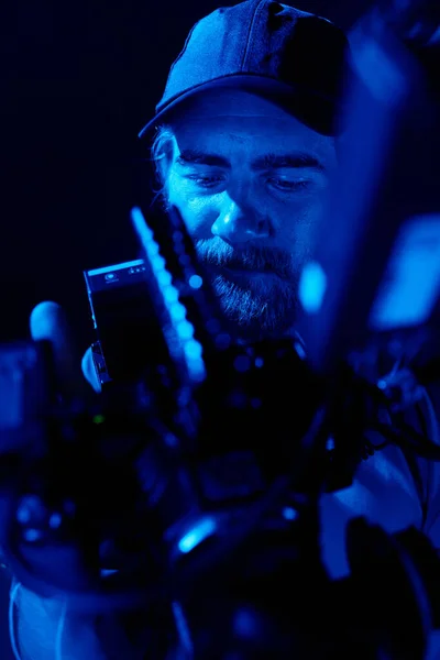 在被蓝光照亮的黑暗工作室拍摄新广告时 留胡子摄影师带着摄像机的面部部分 — 图库照片