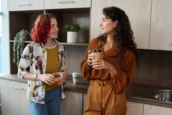两名年轻优雅的女员工穿着时髦的便服 一边站在厨房里一边喝咖啡 一边吃午饭 休息时聊天 — 图库照片