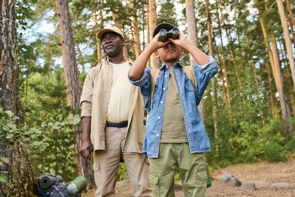 週末のハイキング中に祖父と一緒に松の木の森に立っている間 双眼鏡で見ている犠牲者の男の子 — ストック写真