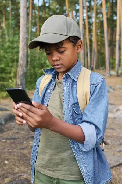 若いですアフリカ系アメリカ人の男の子でスマートフォンのコンサルティングナビゲーターマップながら バックパック旅行中にパス上の深い森の中に立って — ストック写真