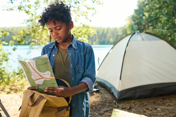 バックパック旅行中にテントや水辺に対してカメラの前に立っている間に地図を見てカジュアルウェアでかわいい前10代の男の子 — ストック写真