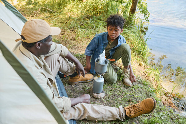 Молодой афроамериканец в повседневной одежде разговаривает со своим дедом, сидя у воды и ожидая, когда будет приготовлена еда.