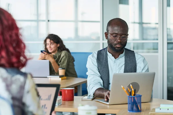 年轻而严肃的非洲裔美国男性分析员坐在笔记本电脑前的桌子旁 用小工具组织反对女同事的工作 — 图库照片