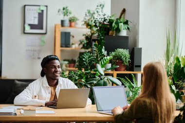 Genç mutlu siyahi adam önlerinde dizüstü bilgisayarı olan sarışın bayan meslektaşlarına bakıyor ve sunum hazırlıyor.