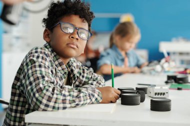 Gözlüklü genç, robotik dersinde öğretmenine bakıyor ve robot parçalarıyla birlikte masanın başında otururken notlar alıyor.